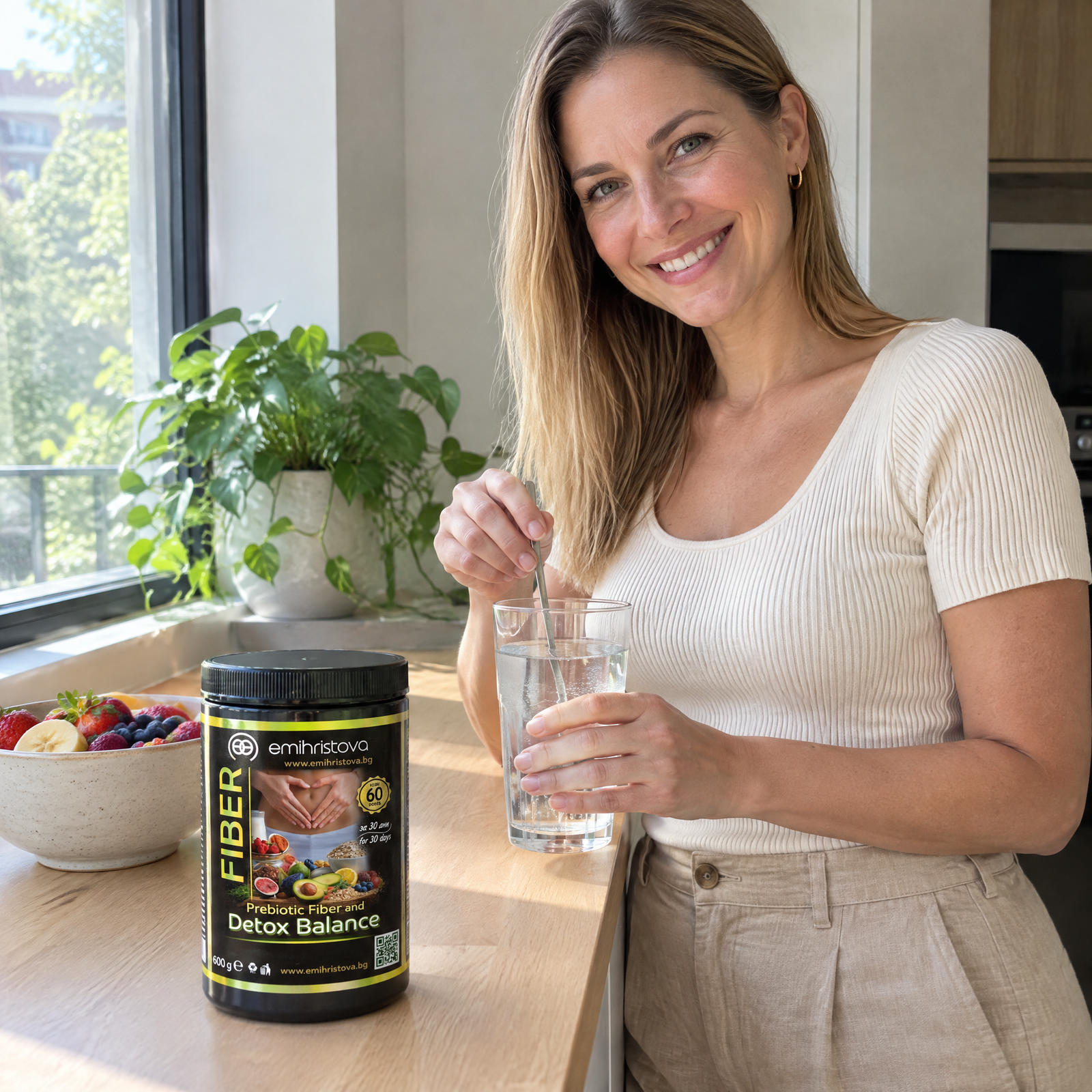 Усмихната жена разбърква чаша вода в кухнята, до нея Detox Balance